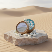 An open round box of Tadpole & Piglet Rock Salt & Driftwood Botanical Wax Melts with blue and white accents rests on a stone slab in the desert, surrounded by hand-poured melts and small white flowers.