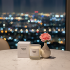 A Tadpole & Piglet Rose Scented Candle with its box rests on a marble table next to a small vase holding a pink carnation, city lights softly blurred through the window in the background.