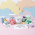 A Mother’s Day scene with pastel gift boxes, a Tadpole & Piglet Rose Scented Candle, paper flowers, and a Happy Mother’s Day card set on a pink surface against a colorful cloud backdrop.