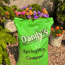 A vibrant green bag of Dandy’s Topsoil & Landscape Supplies Compost Springmix Garden Mulch sits on gravel among blooming marigolds and petunias, with a leafy plant and garden wall behind—ideal for enriching your compost garden mulch.
