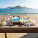 A round box of Tadpole & Piglet’s Seychelles Botanical Wax Melts rests on a wooden table amid pastel flowers, with a scenic ocean view and lush green islands in the background beneath a clear blue sky.