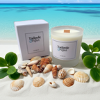 A Tadpole & Piglet Seychelles Scented Candle sits on sandy beach among seashells and leaves, with a turquoise ocean and blue sky in the background.