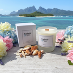 A Tadpole & Piglet Seychelles Scented Candle with an FSC-Certified Wooden Wick sits with its box on wood among seashells and pastel flowers, against a backdrop of a tropical sea and lush green mountains under a blue sky.