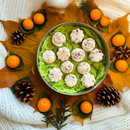 The Tadpole & Piglet Thai Lime & Mango Botanical Wax Melts, shaped like white flowers, are displayed on green leaves with autumn leaves, pinecones, orange fruits, and greenery atop a white knitted fabric.