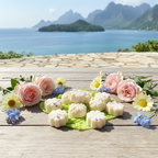 A wooden table featuring Tadpole & Piglet’s Thai Lime & Mango Botanical Wax Melts, shaped like white flowers on green leaves, is adorned with pink roses and daisies, set against a scenic seaside backdrop of mountains and blue water.