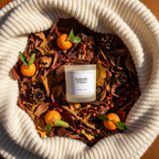 A Tadpole & Piglet Thai Lime & Mango Scented Candle with natural soy wax and an FSC-Certified wooden wick rests on autumn leaves, surrounded by small oranges, pine cones, red berries, and a cozy cream knit blanket.