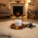 A Tadpole & Piglet Thai Lime & Mango Scented Candle sits on a fluffy white rug with autumn leaves, berries, and a bouquet nearby. String lights and a glowing fireplace add warmth to the cozy living room in the background.