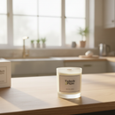 A Tadpole & Piglet Thai Lime & Mango Scented Candle with an FSC-certified wooden wick sits on a wooden kitchen counter in sunlight, with its box and kitchen items softly blurred in the background.