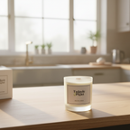 A Tadpole & Piglet Thai Lime & Mango Scented Candle with an FSC-certified wooden wick sits on a wooden kitchen counter in sunlight, with its box and kitchen items softly blurred in the background.