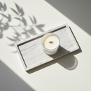 The Tadpole & Piglet Thai Lime & Mango Scented Candle, made with natural soy wax and an FSC-Certified Wooden Wick, rests on a marble tray. Sunlight and leafy shadows set a calm, minimalist tone that highlights the candle’s simple elegance.