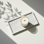The Tadpole & Piglet Thai Lime & Mango Scented Candle, made with natural soy wax and an FSC-Certified Wooden Wick, rests on a marble tray. Sunlight and leafy shadows set a calm, minimalist tone that highlights the candle’s simple elegance.