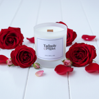 A Tadpole & Piglet Velvet Rose & Oud Scented Candle (Seconds) in a glass jar rests on a white surface, surrounded by red roses, rosebuds, and scattered petals.