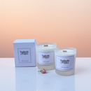 Two white Tadpole & Piglet Velvet Rose & Oud Scented Candle (Seconds) in glass jars sit beside a matching box and a pink flower bud on a soft gradient background.