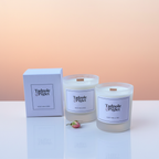 Two white Tadpole & Piglet Velvet Rose & Oud Scented Candle (Seconds) in glass jars sit beside a matching box and a pink flower bud on a soft gradient background.