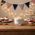 A Tadpole & Piglet Velvet Rose & Oud Scented Candle (Seconds) glows on a wooden table with pretzels, cupcakes, and cookies, while string lights and red, white, and blue bunting set a festive mood in the background.