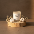 A Tadpole & Piglet Wild Fig & Cassis Scented Candle with a wooden wick rests on a round wooden platform, surrounded by baby's breath and cotton flowers, as warm natural light creates soft shadows.