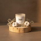A Tadpole & Piglet Wild Fig & Cassis Scented Candle with a wooden wick rests on a round wooden platform, surrounded by baby's breath and cotton flowers, as warm natural light creates soft shadows.