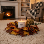 A Tadpole & Piglet Wild Fig & Cassis Scented Candle with a wooden wick glows in a glass holder, centered in an autumn leaf wreath on the carpet, as a cozy fireplace warms the inviting living room in the background.