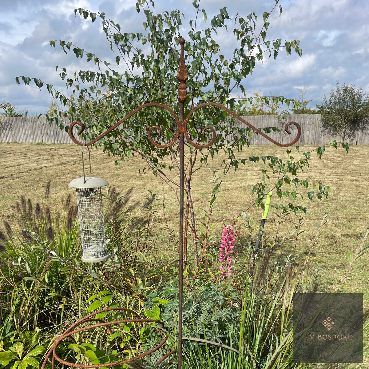 Raw steel bird feeding stake with a hanging seed feeder beside a young tree