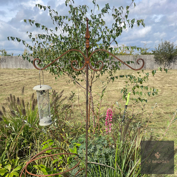 Raw steel bird feeding stake with a hanging seed feeder beside a young tree