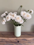 A Bishy Barnabees Cottage Garden "Cornflower Snowman" vase, filled with fluffy white double blooms, sits on a wooden surface against a plain light wall.