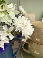 Cornflower Snowman by Bishy Barnabees Cottage Garden features white and purple double blooms in a blue vase, displayed beside stacked cream ceramic cups and saucers against a light background.