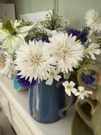 The Cornflower Snowman ceramic mug by Bishy Barnabees Cottage Garden, filled with white and purple cottage-garden blooms, rests on a white surface beside a green heart-shaped ceramic piece.