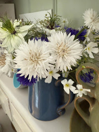 The Cornflower Snowman ceramic mug by Bishy Barnabees Cottage Garden, filled with white and purple cottage-garden blooms, rests on a white surface beside a green heart-shaped ceramic piece.