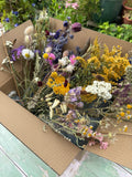 The Bishy Barnabees Cottage Garden Dried Flower Craft Box, filled with a vibrant mix of yellow, purple, white, and pink blooms for crafts, sits on a weathered green surface against a garden backdrop.