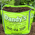 A large, bright green Dandy's Topsoil & Landscape Supplies bag labeled "Manure Farmyard" filled with soil sits on gravel in a lush garden, advertising turf, topsoil, spent mushroom compost, phone number, and website.