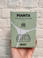 Hand holding a Grow Gang Pianta Full Spectrum Plant Light Bulb box against a white brick wall