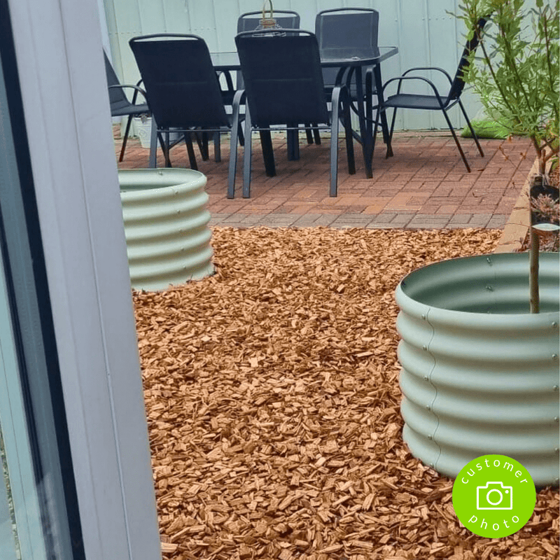 A backyard patio with a table and six chairs on pavers is bordered by Dandy's Topsoil & Landscape Supplies Hardwood Path Chips from a bulk bag, featuring two round raised garden beds and a partially visible sliding glass door in the foreground.