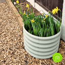 A round, pale green metal planter with yellow daffodils sits on Dandy's Topsoil & Landscape Supplies Hardwood Path Chips beside a garden bed and wooden fence. A green customer photo badge appears in the bottom right corner.