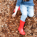 A child, visible from the waist down in blue jeans, a blue shirt, and bright red rain boots, kneels on Dandy's Topsoil & Landscape Supplies' Hardwood Path Chips—an ideal chicken run base from a bulk bag.