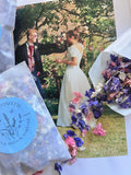 A bride and groom smile outdoors with a bouquet, as Bishy Barnabees Cottage Garden's Larkspur Confetti (Mixed Colours) 4g—biodegradable petals in pink and purple—spills from blue packets across the photo.