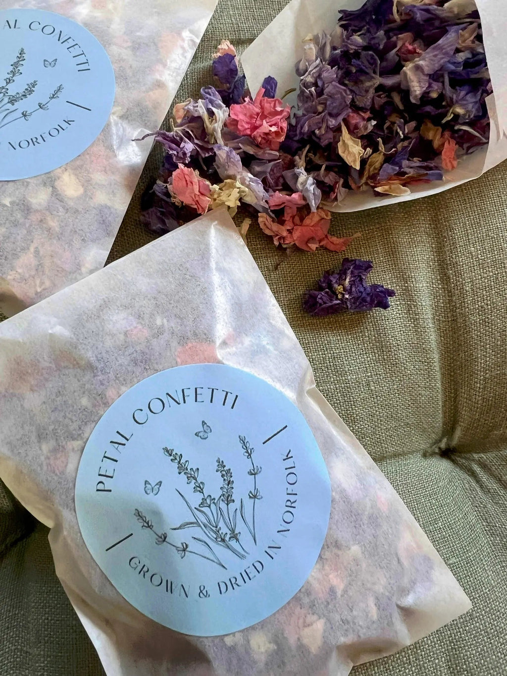 Larkspur Confetti (Mixed Colours) 4g – paper packets on beige fabric, one open showing petals