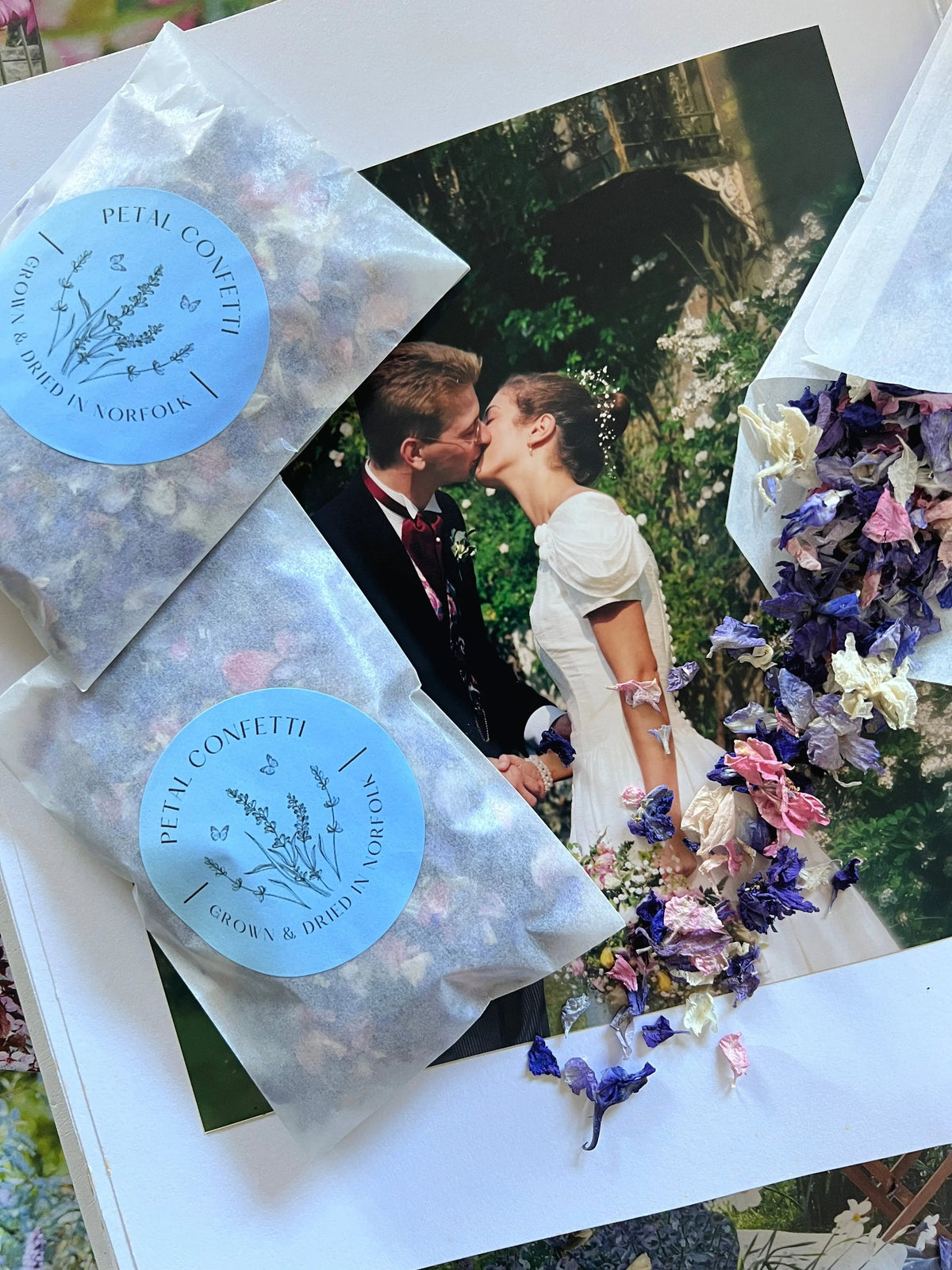 Larkspur Confetti packets on an open book with a bride and groom, pastel petals scattered