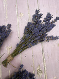 The Lavender Dried (Small) from Bishy Barnabees Cottage Garden Ltd is tied with twine, elegantly displayed on a rustic, light purple wooden surface—ideal as a natural room fragrance.