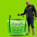A smiling person in a black Dandy’s jacket and beige boots stands beside a large green bag of Dandy's Topsoil & Landscape Supplies Compost Peat Free Multi Purpose Welsh, set against a vivid green background. The bag shows the company name, contact info, and website.