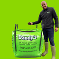 A smiling man in boots and a Dandy’s Topsoil & Landscape Supplies jacket stands by a large, bright green Dandy’s bag filled with Topsoil Welsh Multi Purpose, featuring company contact details on a solid green background.