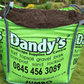 A large green bag labeled Dandy's Topsoil & Landscape Supplies sits on grass, filled with dark soil. It advertises Topsoil Welsh Multi Purpose, turf, gravel, bark, organic compost, sand, rock salt, website and contact details.