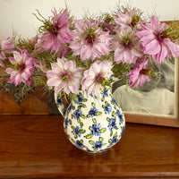 A ceramic jug from Bishy Barnabees Cottage Garden, featuring Nigella Mulberry Rose (Love-in-a-mist) with pale pink blooms and feathery green foliage, sits on a wooden surface next to a black-and-white photo in a frame.