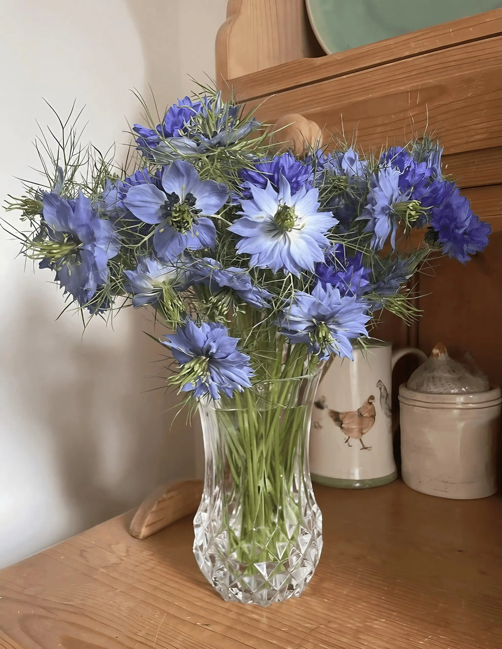 Blue and purple Nigella Miss Jekyll Mixed flowers in a faceted glass vase on a wooden worktop