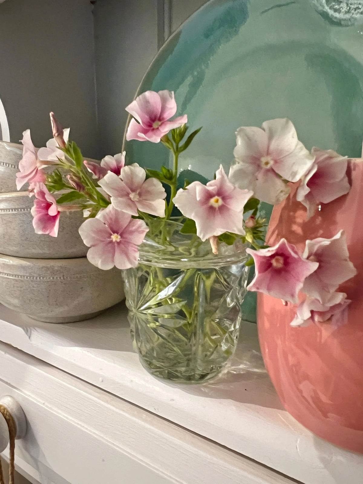 Glass jar of pale pink and white Phlox Blushing Bride beside bowls and a coral vase