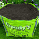 A large green Bulk Bag of Dandy’s Topsoil & Landscape Supplies Compost Peat Free Multi Purpose Welsh sits on garden grass filled with dark topsoil, while flowers bloom in the background.