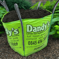 A large green bag labeled "Soil Improver Conditioner" from Dandys Landscape Supplies sits on a garden path, filled with dark soil improver and surrounded by lush plants and greenery.