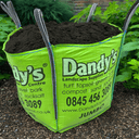 A large green "Dandy's Topsoil & Landscape Supplies" bag filled with dark Compost Peat Free Multi Purpose Welsh sits on gravel near green plants. The bag promotes topsoil, gravel, bark, and compost, plus contact details.