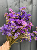 A hand holds a bouquet of Statice Dried flowers by Bishy Barnabees Cottage Garden in front of a gray wooden wall.