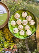 A round box from Tadpole & Piglet, filled with green tissue paper, displays ten white Thai Lime & Mango Botanical Wax Melts shaped like flowers and domes, set on a floral surface that evokes a fresh garden atmosphere.
