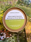 A round Tadpole & Piglet container labeled "Thai Lime & Mango Botanical Wax Melts" rests on colorful garden cards; the label features hand-poured wax melts with lime, mango, peach, and grapefruit scents.